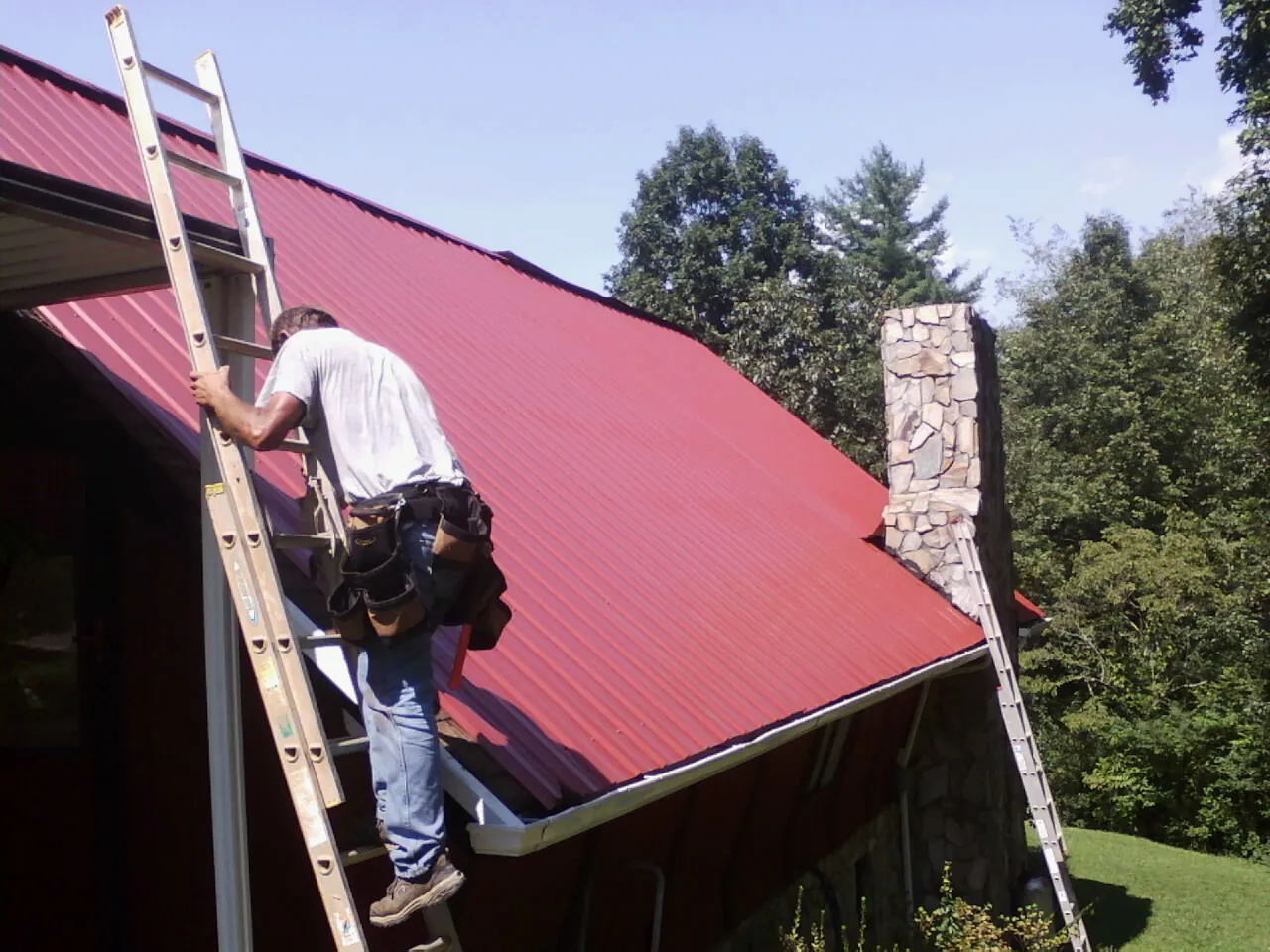 Roofer on a ladder beside a red metal roof and stone chimney — Soffit & Fascia Repair by Roofing Brothers in Walters